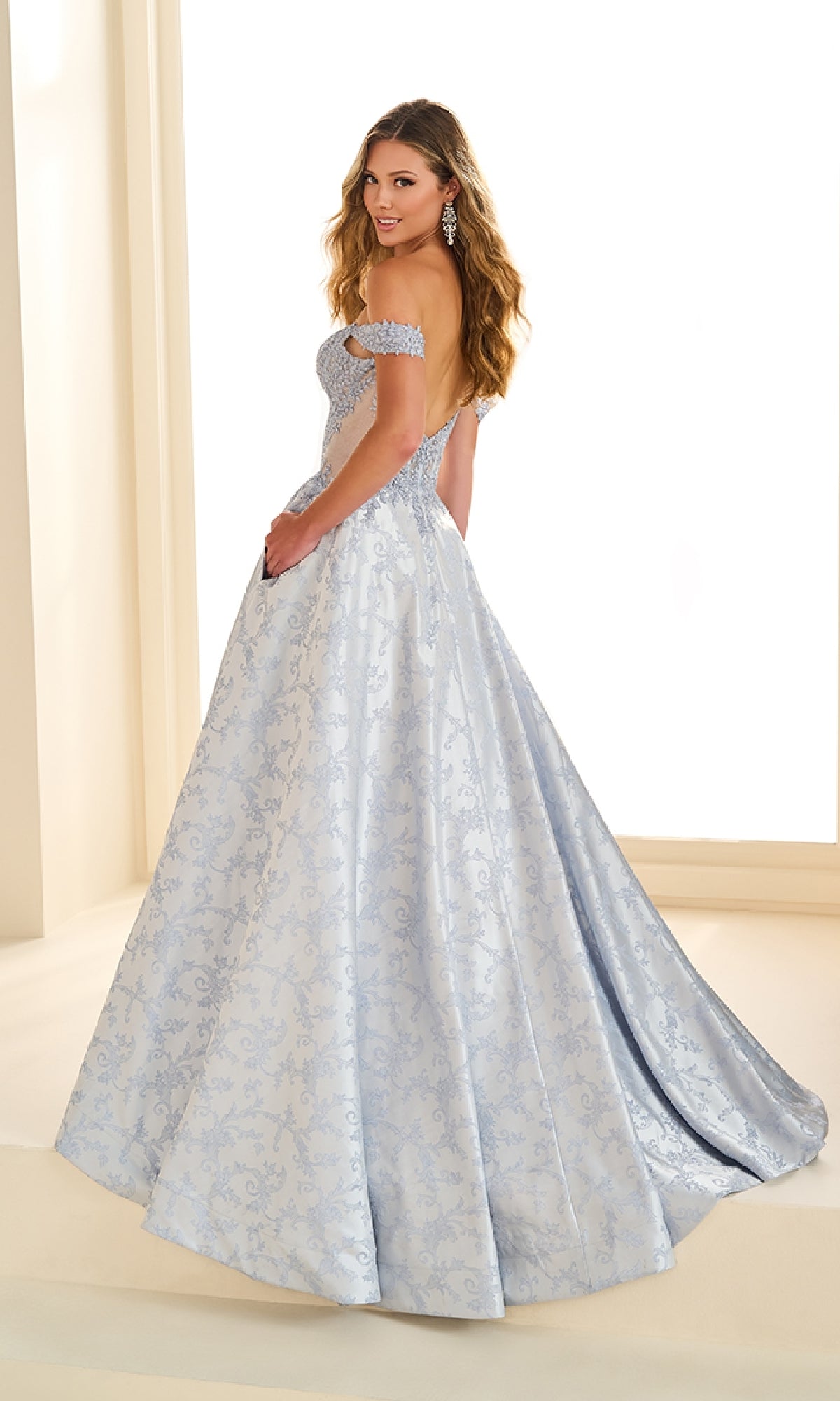A woman wearing the Ellie Wilde EW36078 long prom dress with a light blue embellished corset bodice and a patterned skirt stands facing backward, looking over her shoulder and smiling.